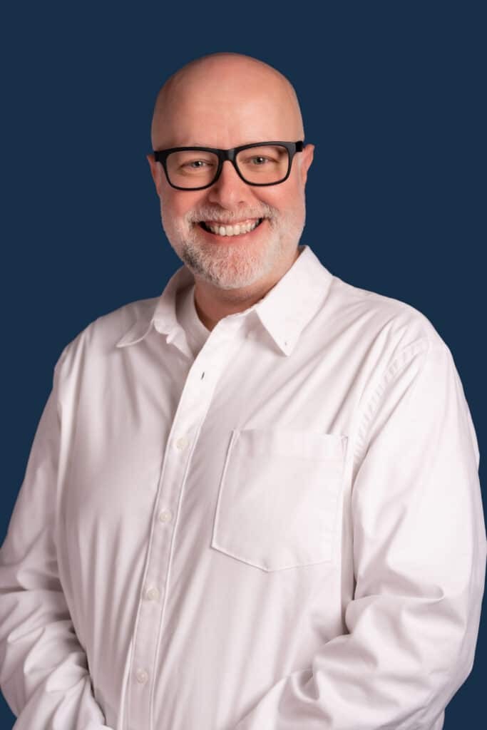A smiling bald man with a beard and glasses in a white button-up shirt stands against a solid dark blue background.