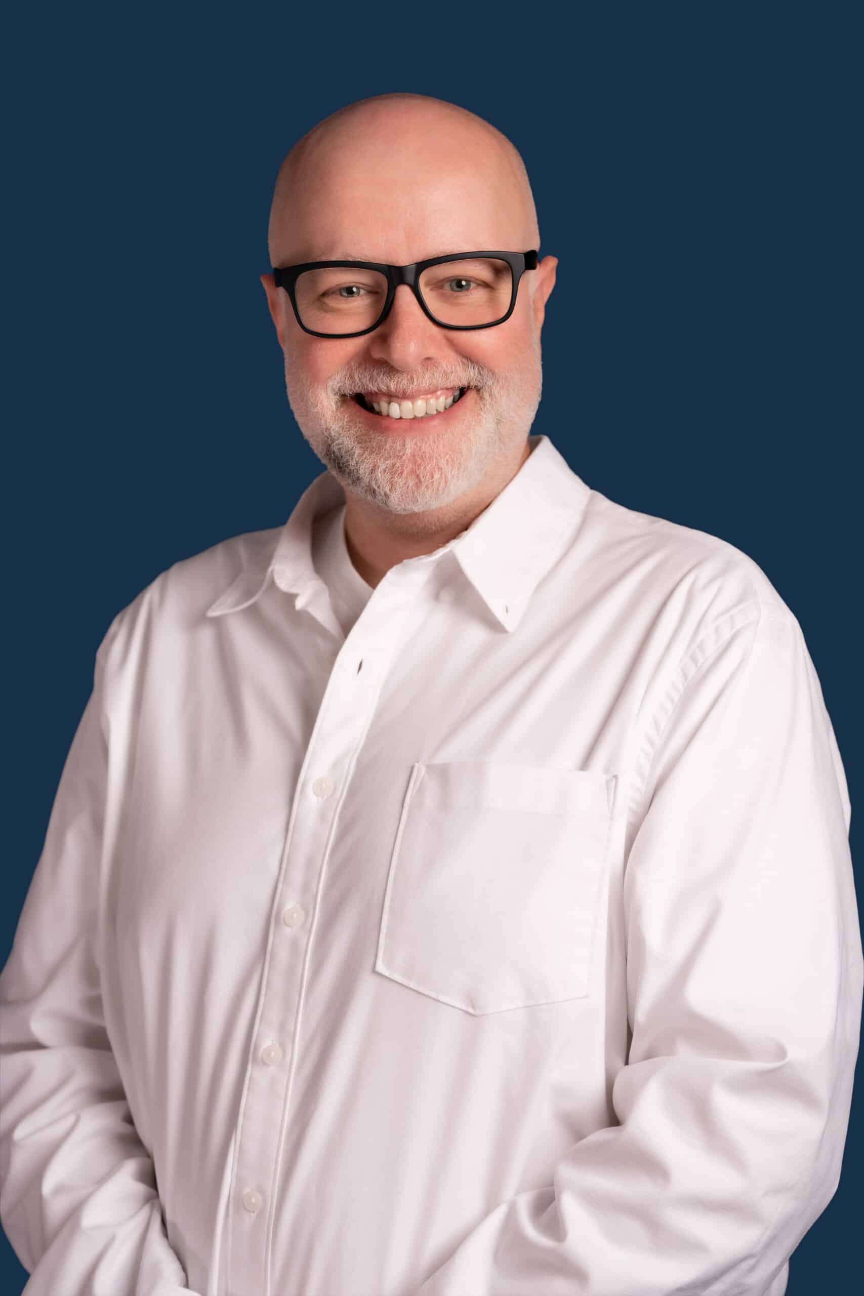 A smiling bald man with a beard and glasses in a white button-up shirt stands against a solid dark blue background.