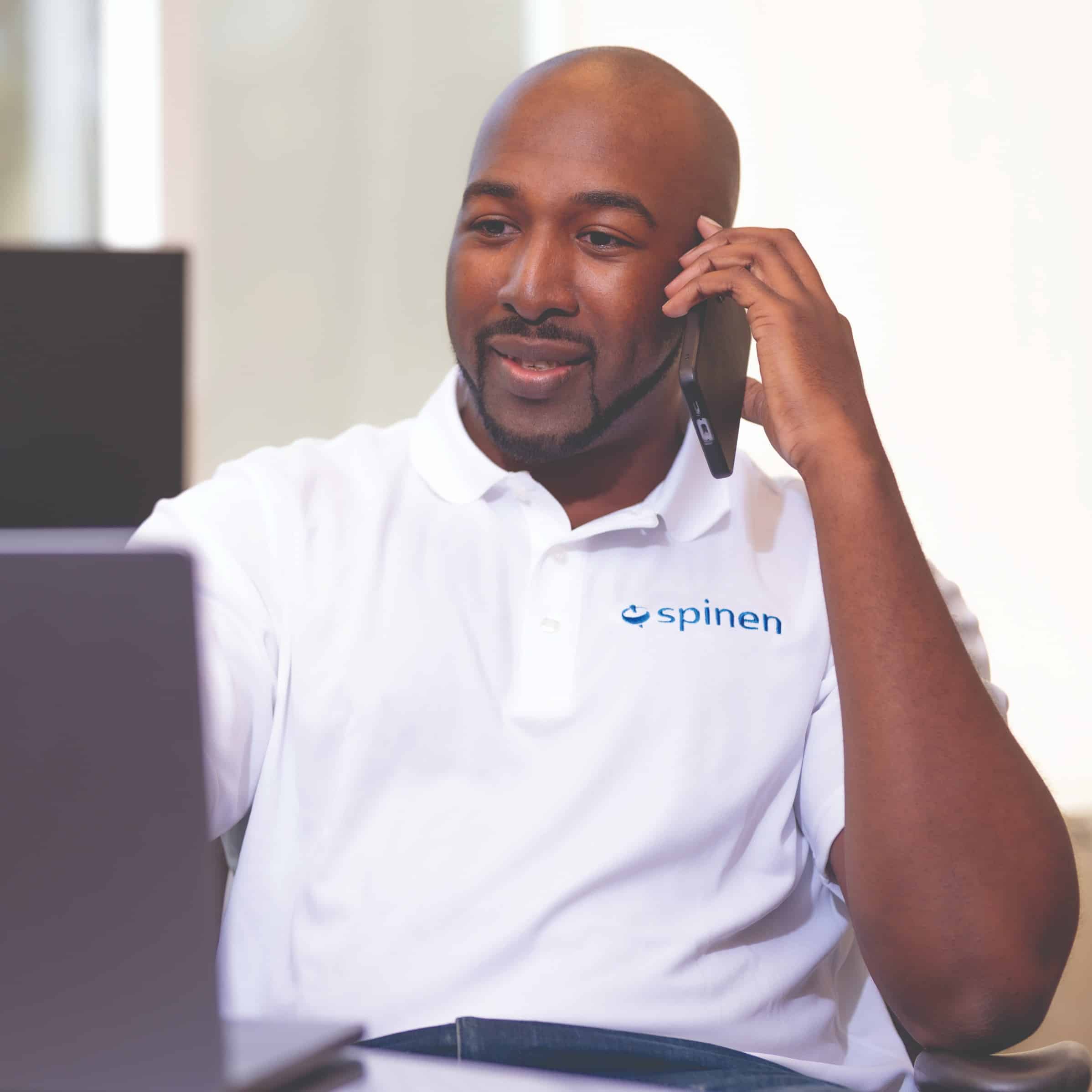 A man wearing a white spinen polo shirt sits at a desk, talking on the phone and looking at a laptop, with a blurred office background.