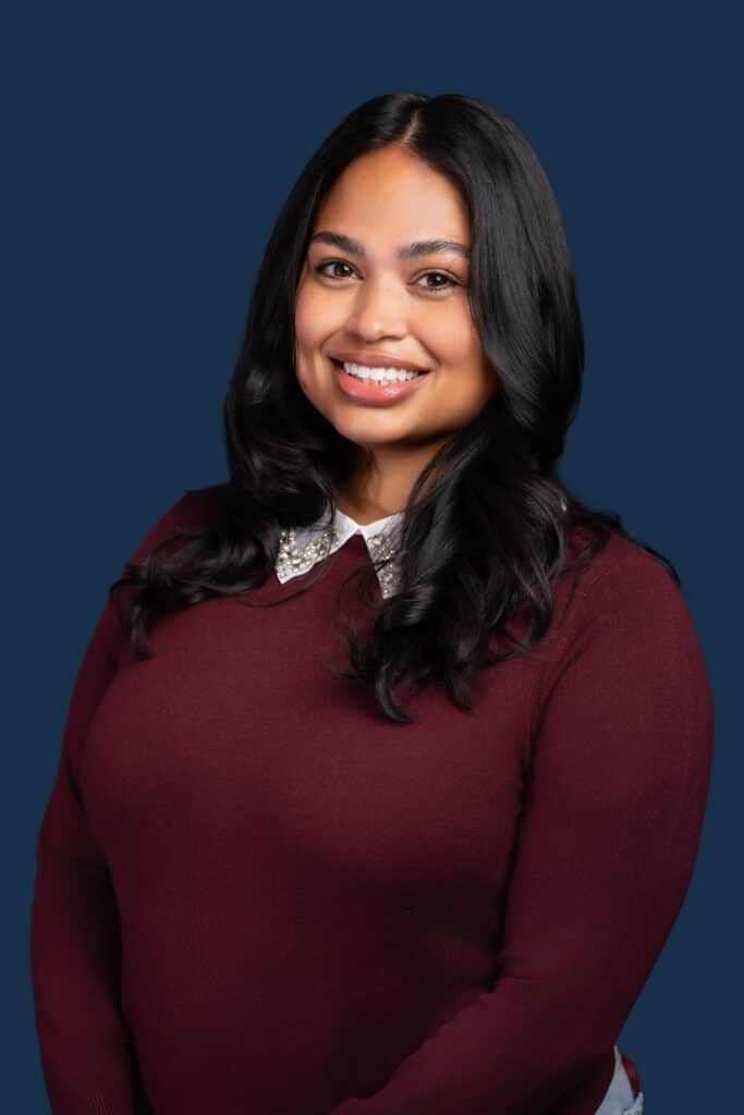 A woman with long dark hair in a maroon sweater over a white collared shirt smiles against a solid dark blue background.