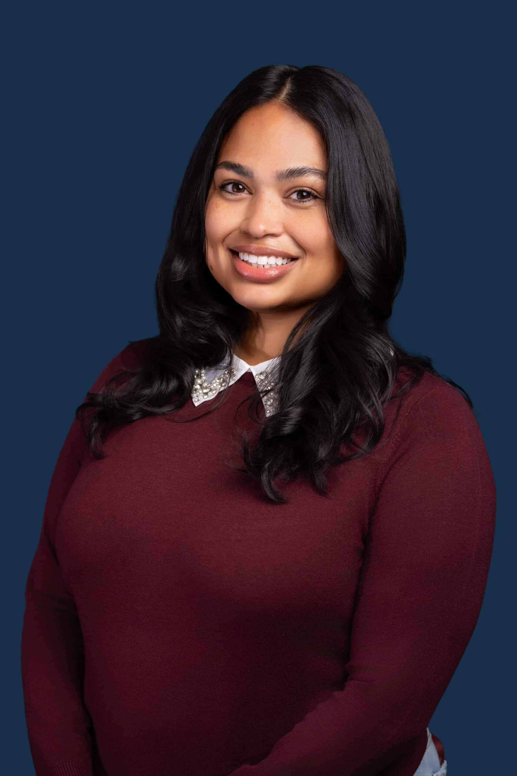 A woman with long dark hair in a maroon sweater over a white collared shirt smiles against a solid dark blue background.