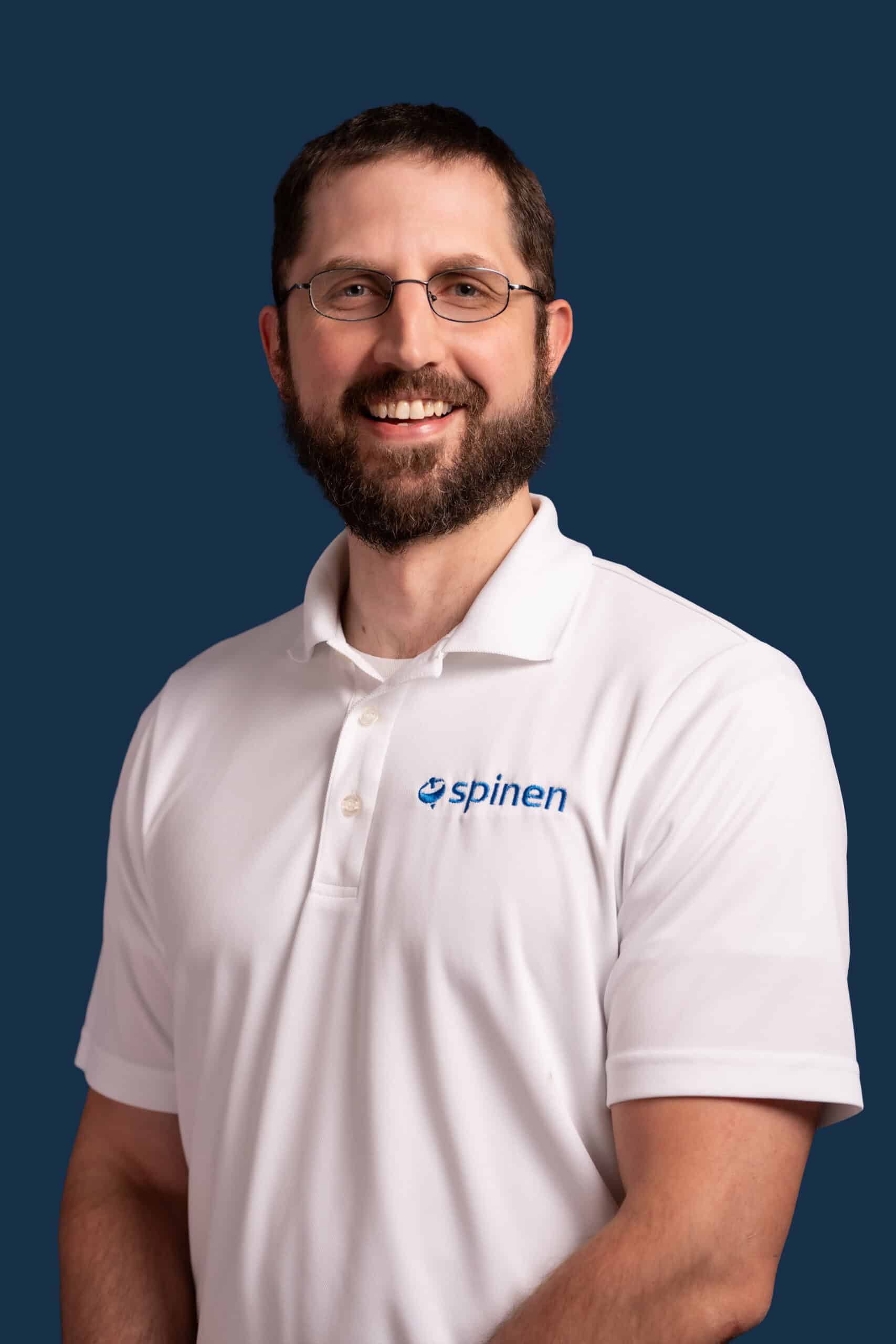 A man with a beard and glasses smiles, wearing a white polo shirt with the spinen logo, standing against a solid dark blue background.