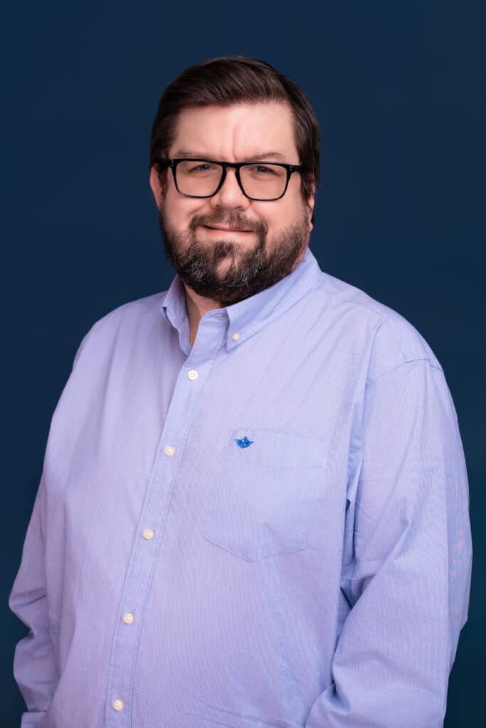 A man with short dark hair, a beard, and glasses in a light blue button-up shirt stands against a dark blue background, smiling.