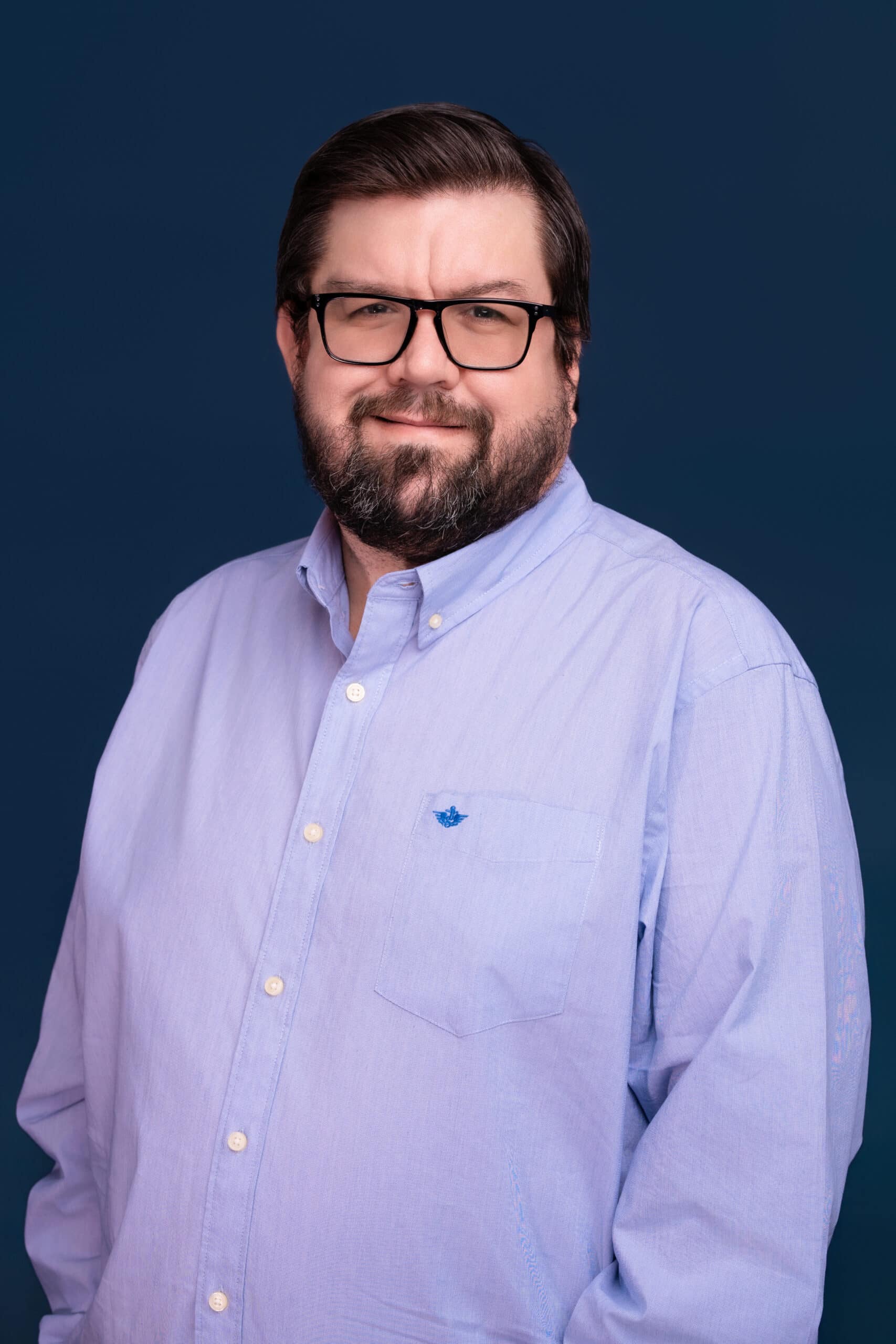 A man with short dark hair, a beard, and glasses in a light blue button-up shirt stands against a dark blue background, smiling.