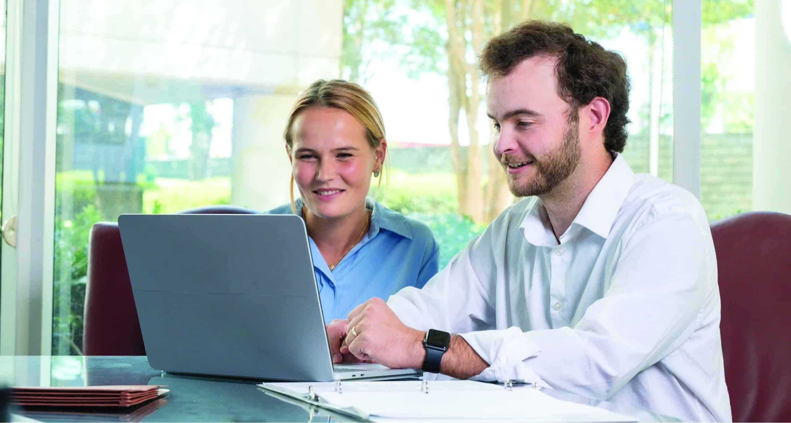 Two people smile at a laptop across a table with papers; large windows let in light and reveal greenery outside.