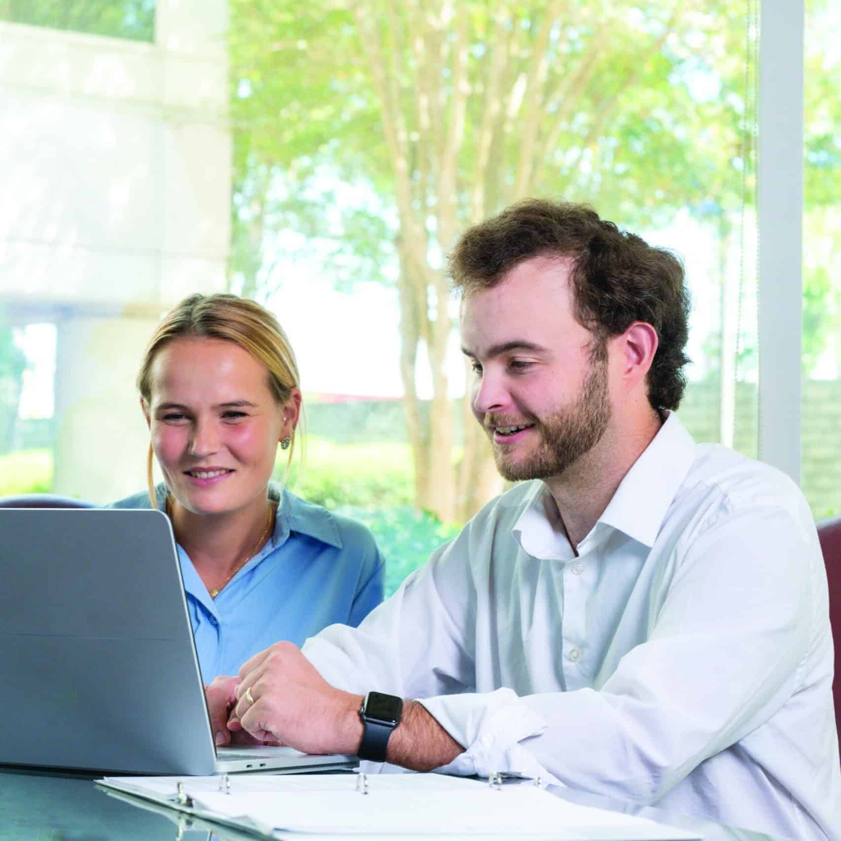 A man and woman smile at a laptop together at a sunlit table with papers and a binder. Trees are visible through big windows.