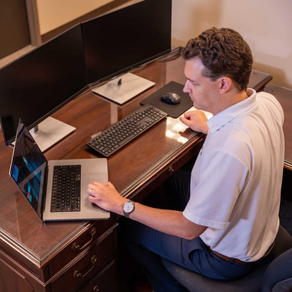 A man in a white shirt sits at a wooden desk working on a laptop, with two monitors, keyboard, and mouse. He looks focused.
