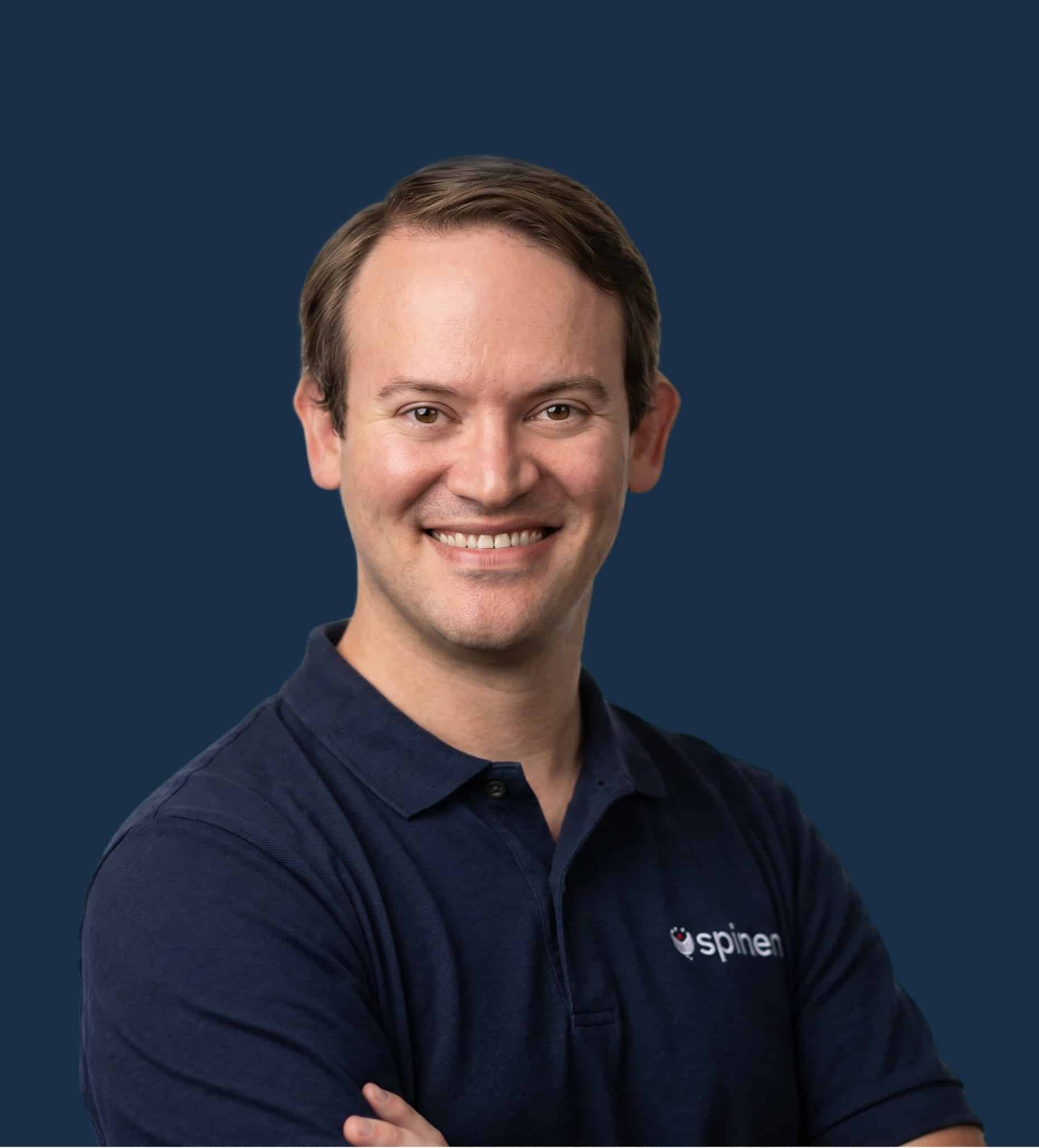 Smiling man with short brown hair wearing a navy polo shirt with a spinen logo, standing against a plain dark blue background, arms crossed.