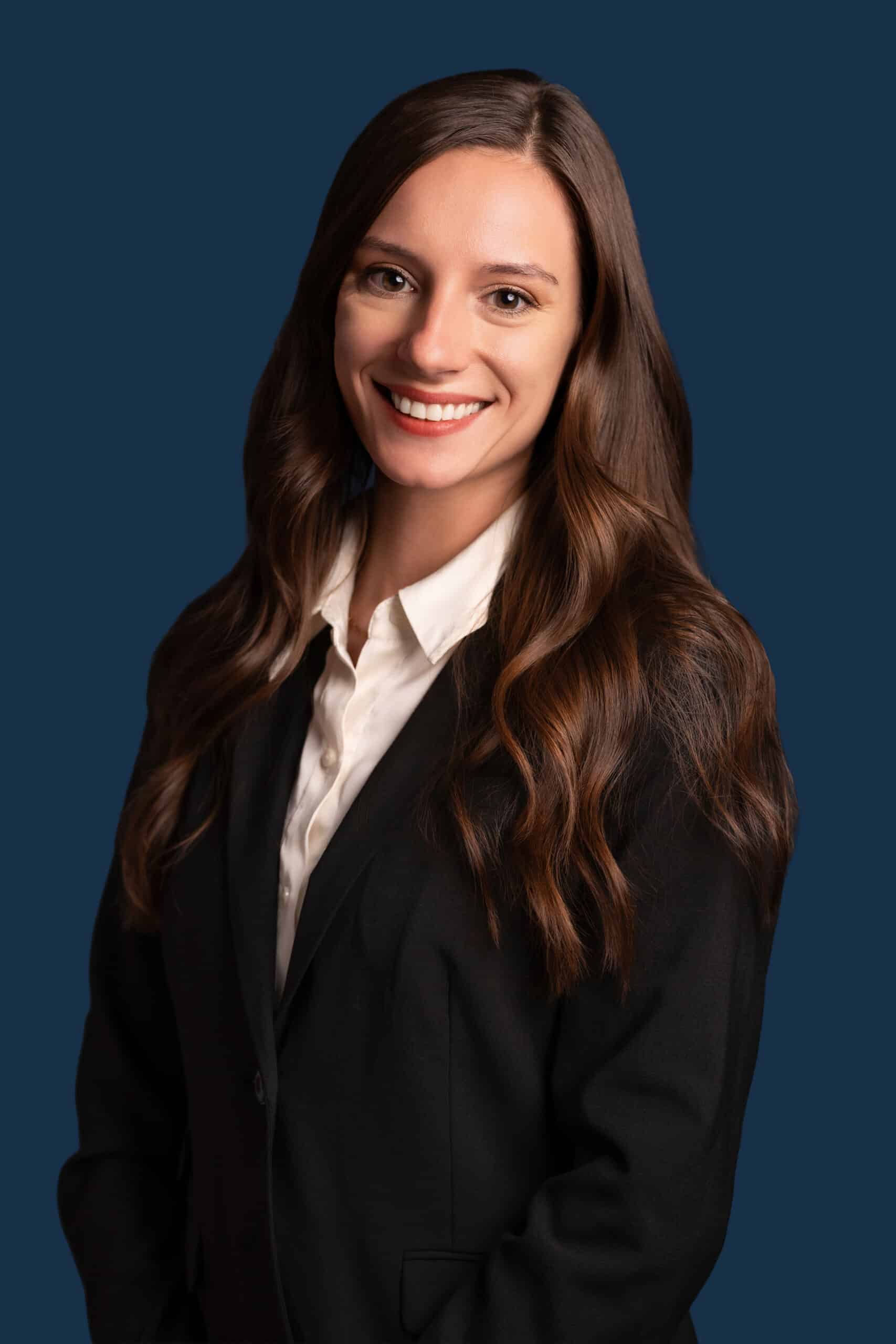 A woman with long brown hair in a black blazer and white blouse smiles at the camera against a solid dark blue background.