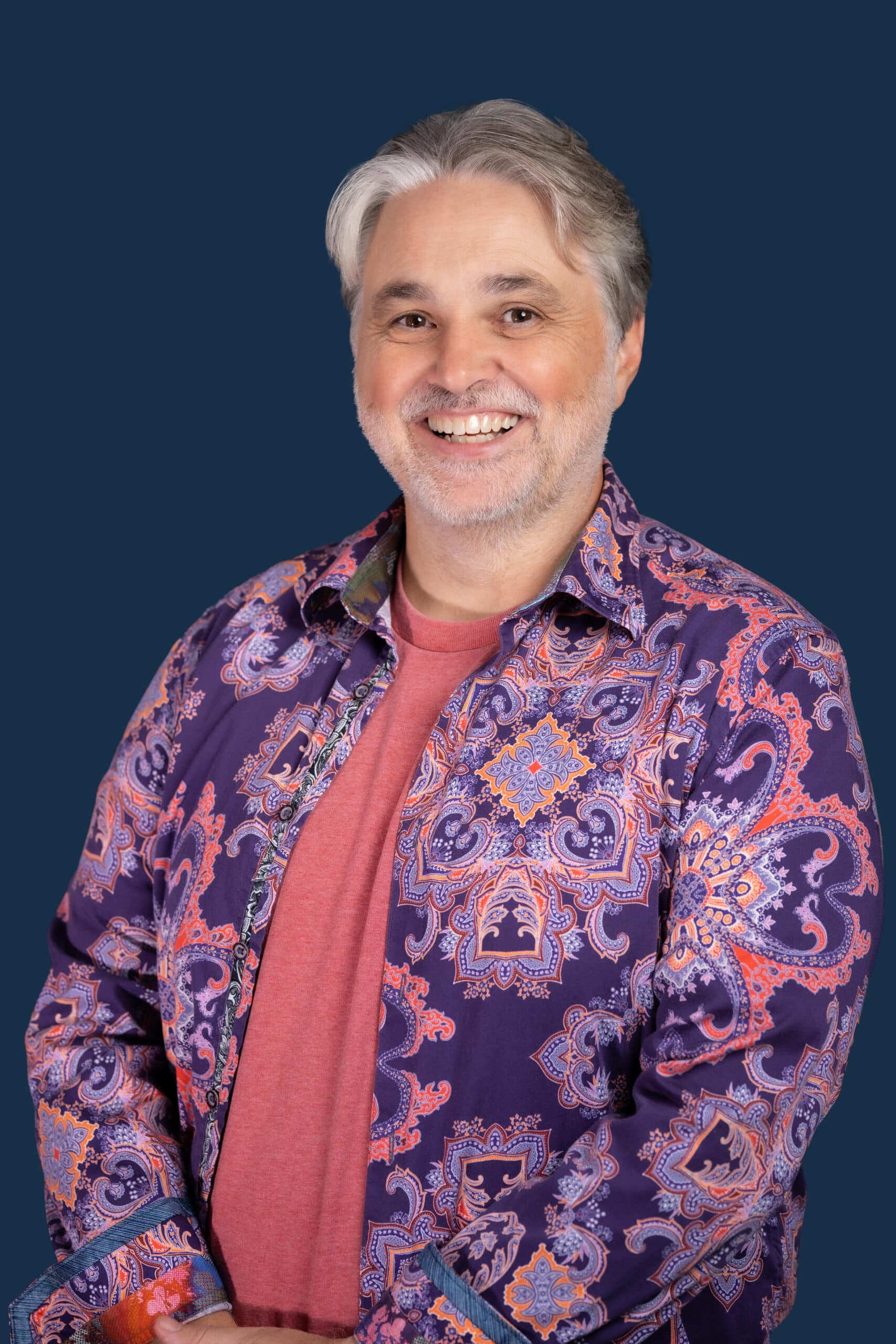 A smiling middle-aged man with gray hair and a beard in a colorful paisley shirt over a red tee stands before a dark blue backdrop.