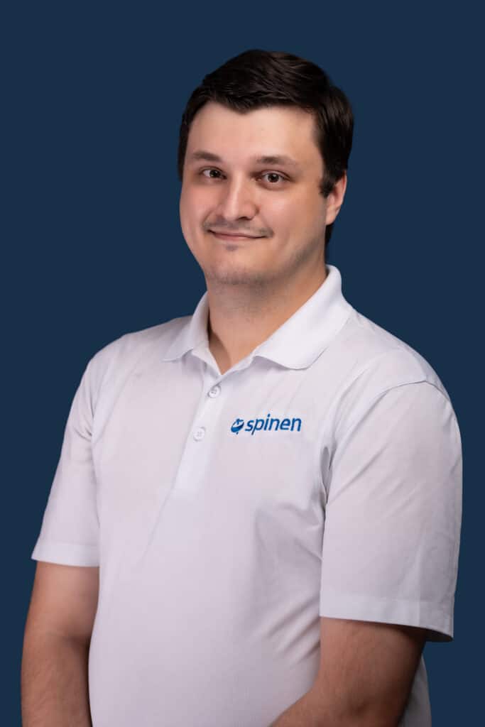 A man with short dark hair wears a white polo shirt with a blue spinen logo, standing against a plain dark blue background, smiling softly.