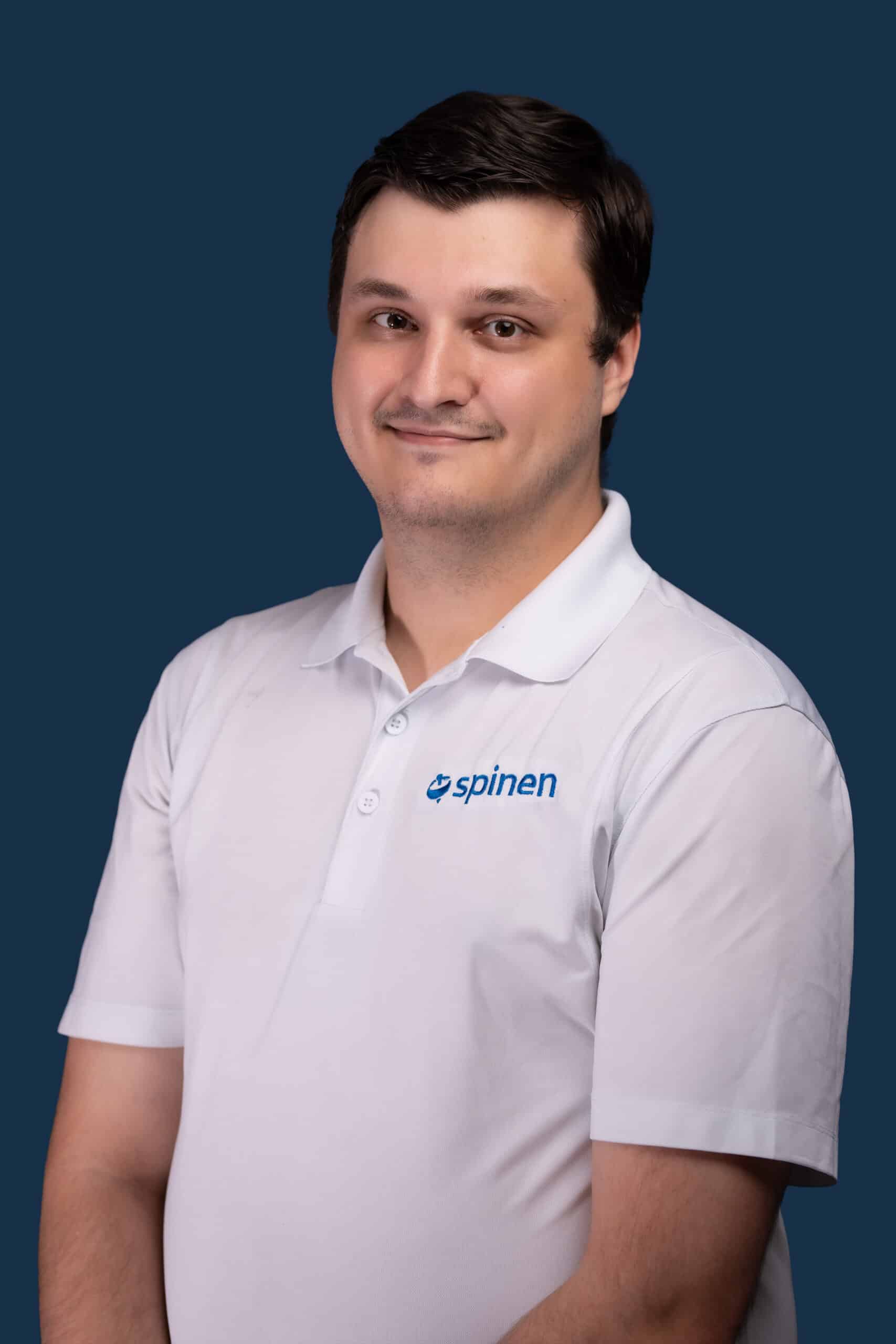 A man with short dark hair wears a white polo shirt with a blue spinen logo, standing against a plain dark blue background, smiling softly.