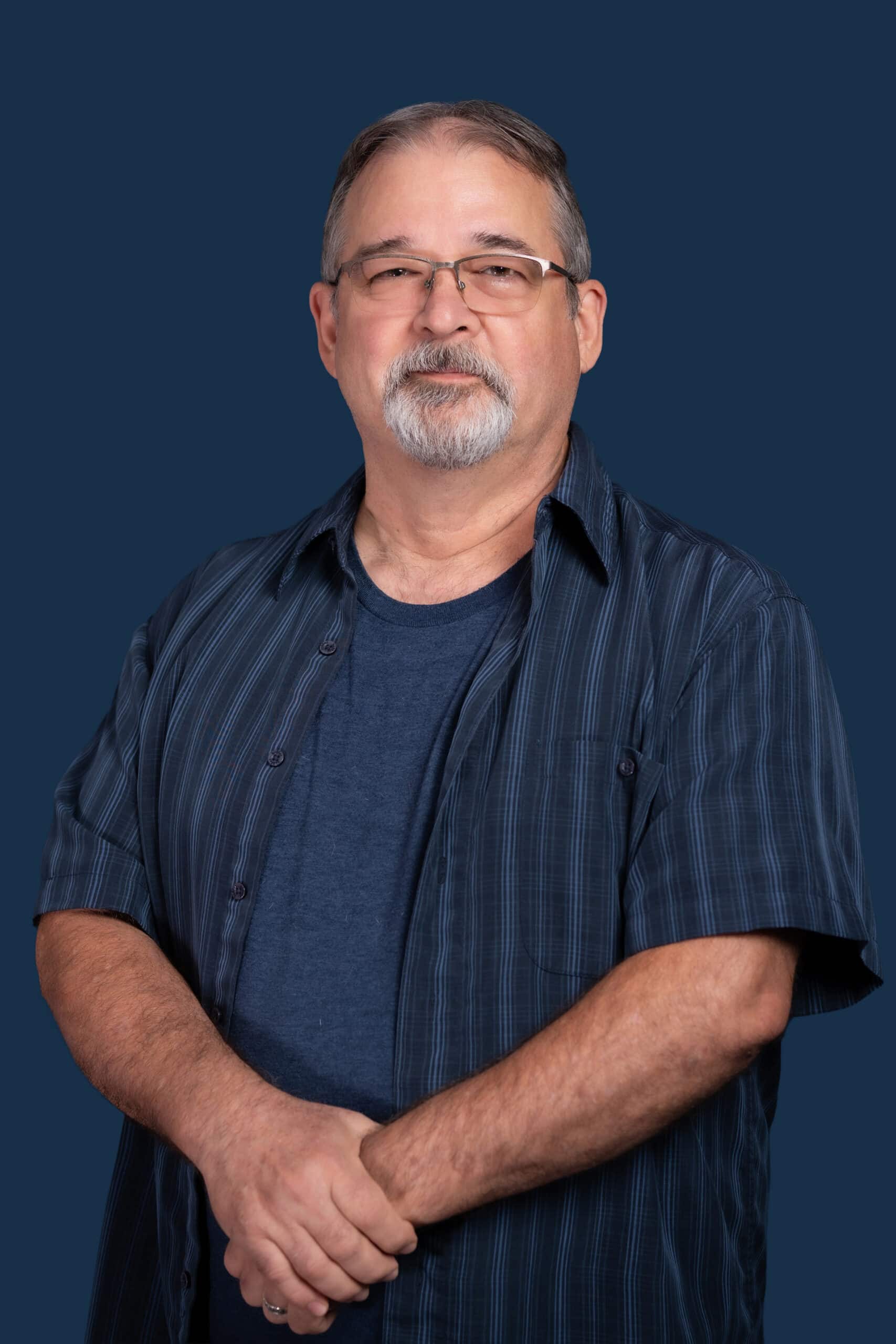 Middle-aged man with gray hair, mustache, and glasses wears navy t-shirt and striped shirt, stands before a dark blue background.