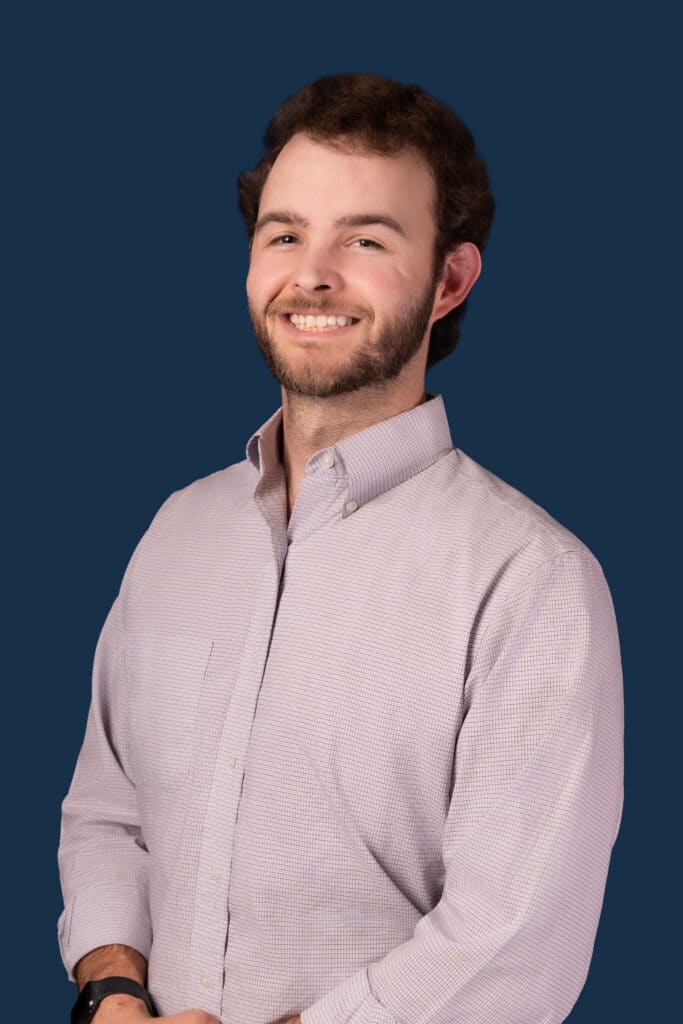 A man with short brown hair and a beard smiles in front of a solid dark blue background, wearing a light long-sleeve shirt with sleeves rolled up.