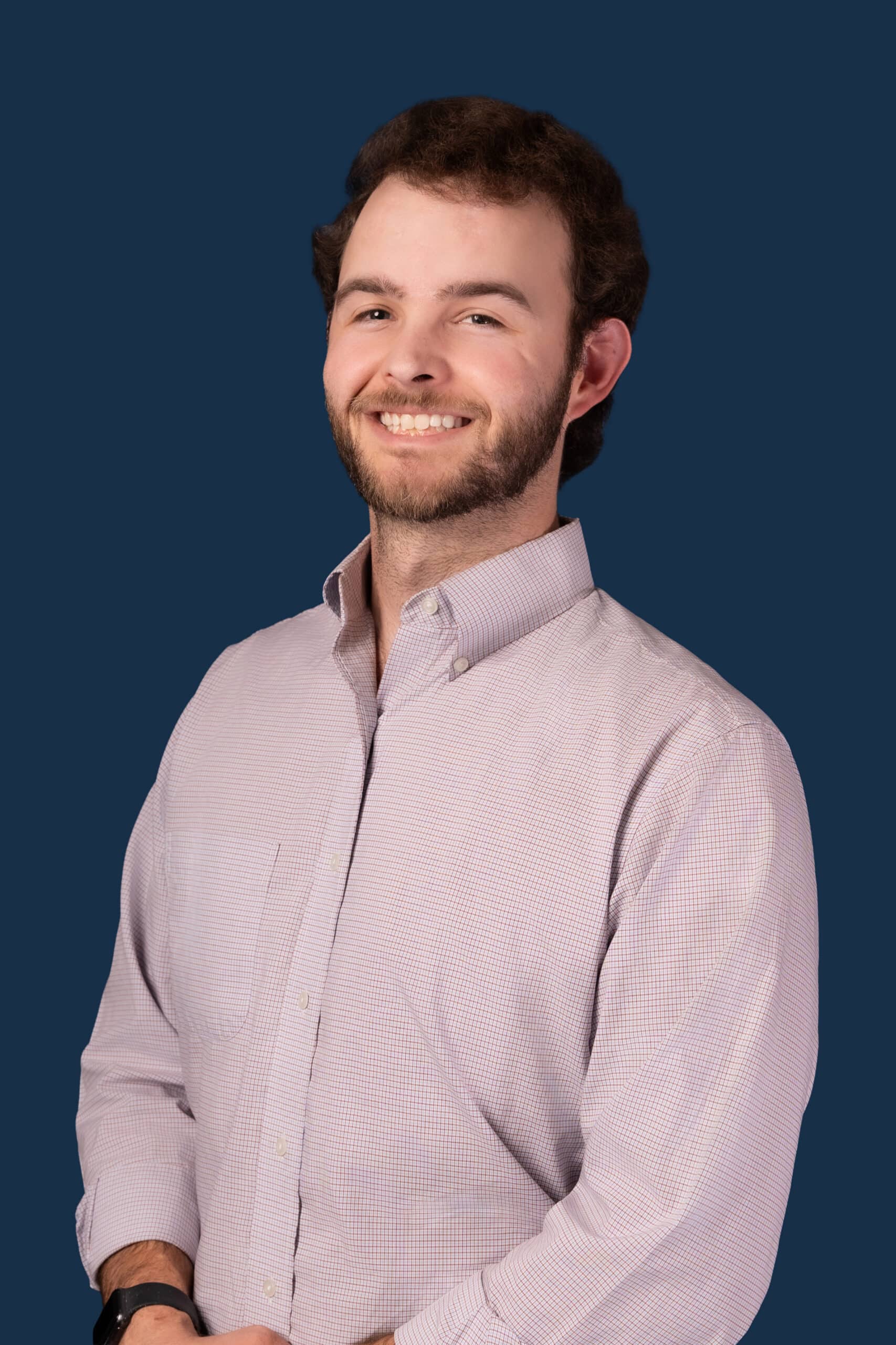A man with short brown hair and a beard smiles in front of a solid dark blue background, wearing a light long-sleeve shirt with sleeves rolled up.