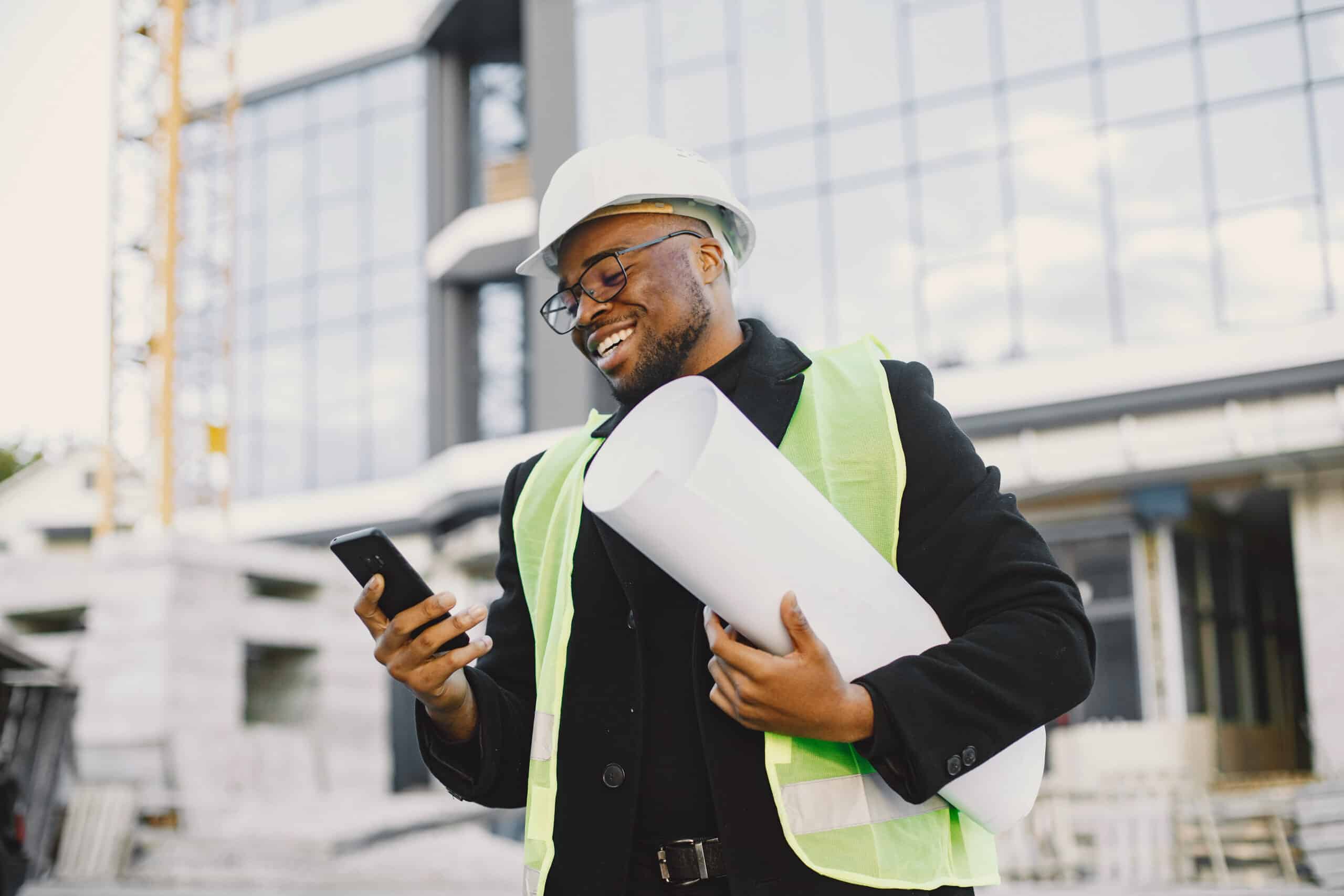 A smiling construction worker in a hard hat and safety vest holds blueprints and checks a smartphone by a glass building under construction.