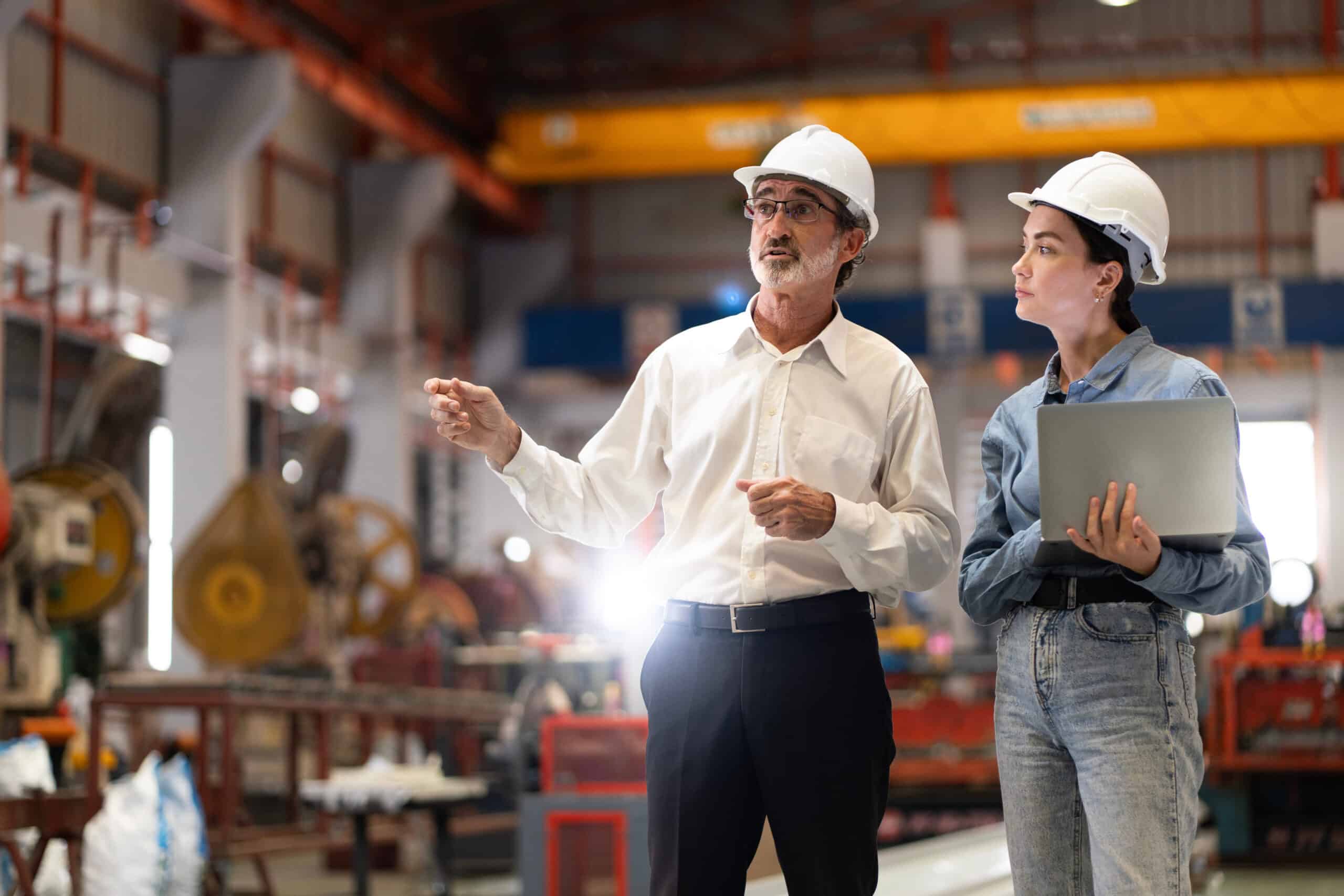 Two engineers in safety helmets stand in a factory; the older man gestures as the younger woman listens with a laptop, machinery behind them.