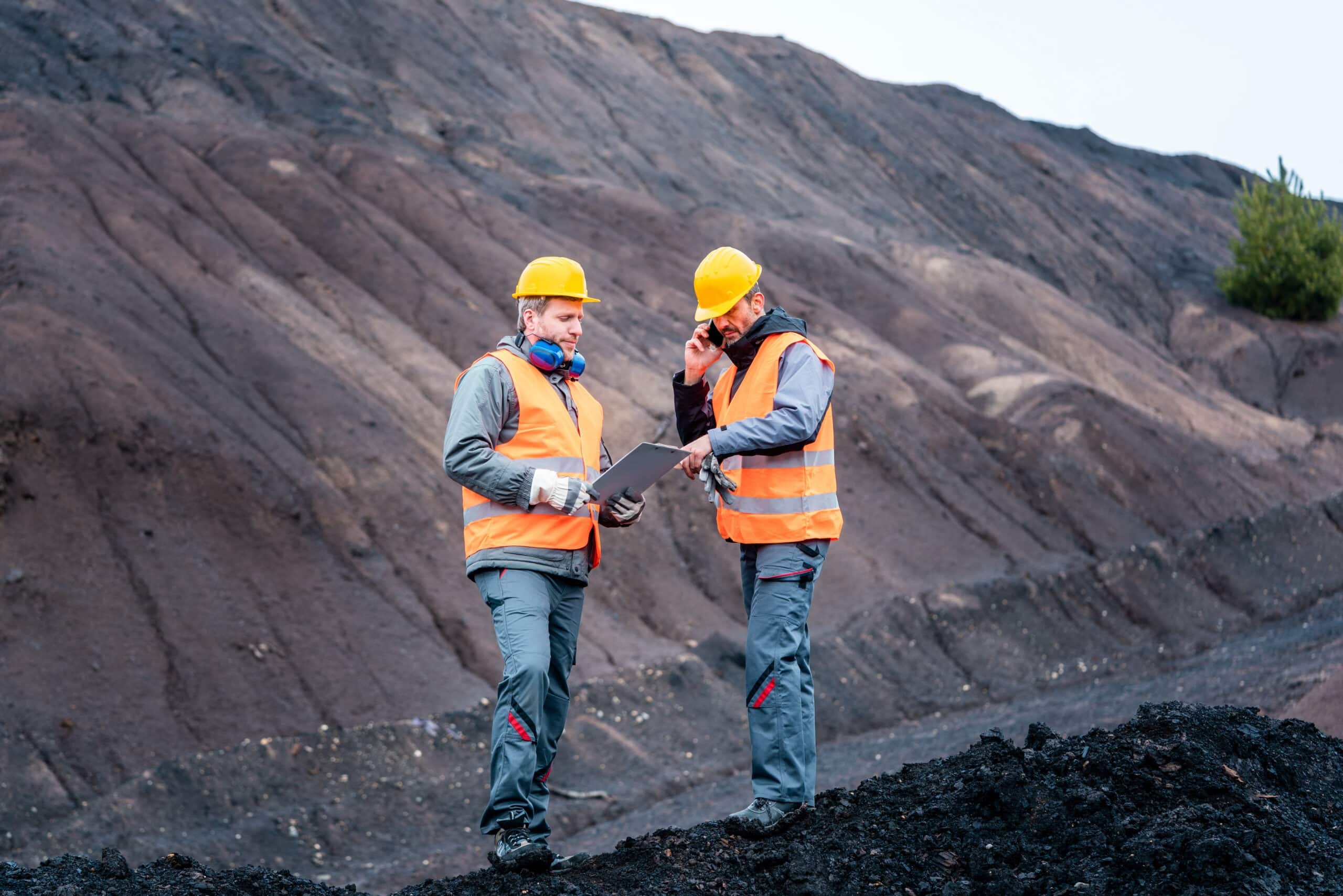 Two workers in orange safety vests and yellow helmets at a rocky mining site; one holds a clipboard, the other uses a radio.