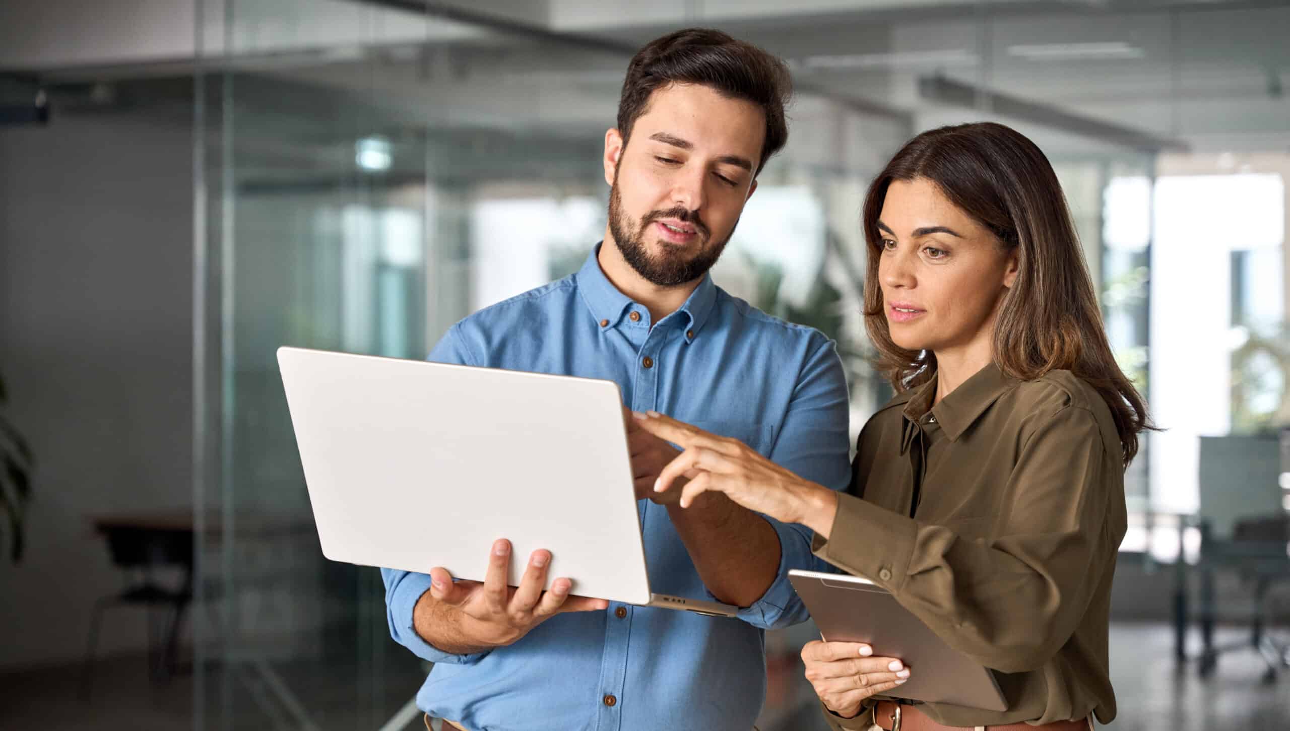 Two people in a modern office look at a laptop together; the man points at the screen while the woman with a tablet listens closely.