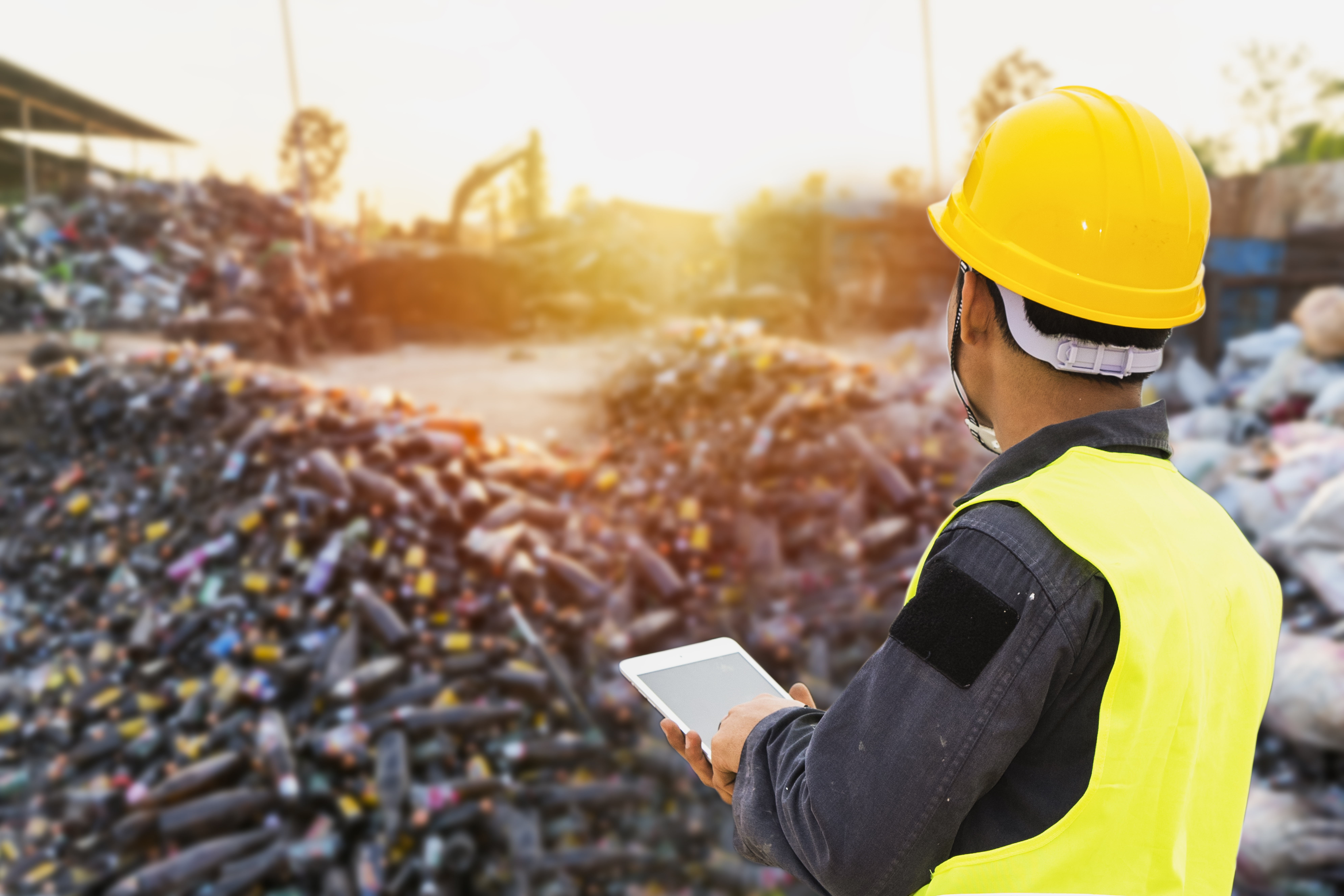 A worker in a yellow safety vest and hard hat uses a tablet while inspecting a large pile of bottles and waste at a recycling facility.