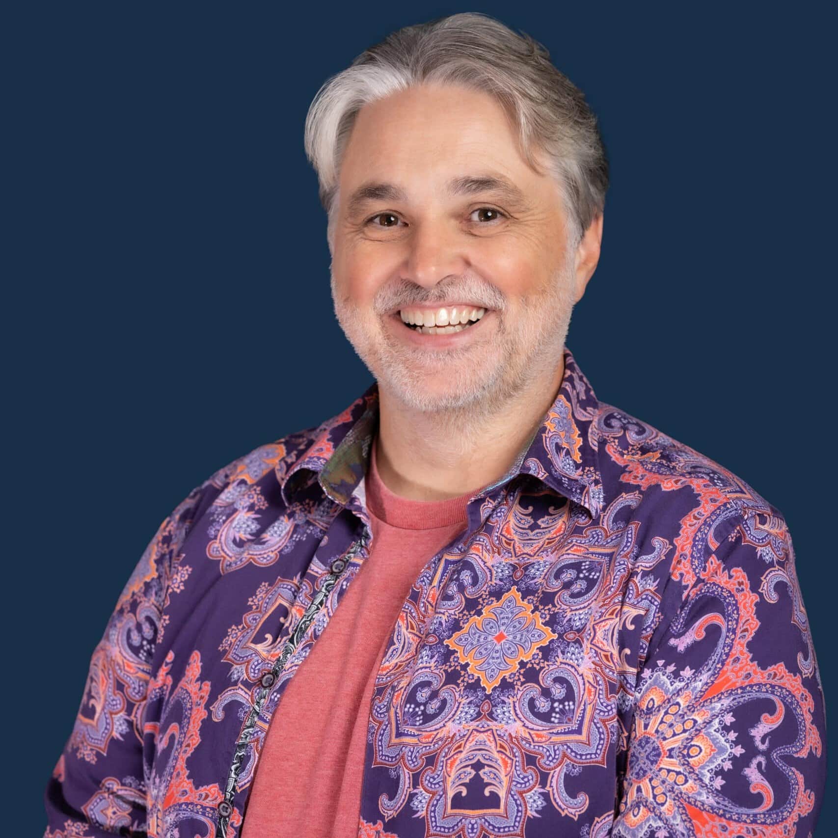 A smiling middle-aged man with gray hair and a short beard wears a vibrant paisley shirt over a red t-shirt, against a dark blue background.
