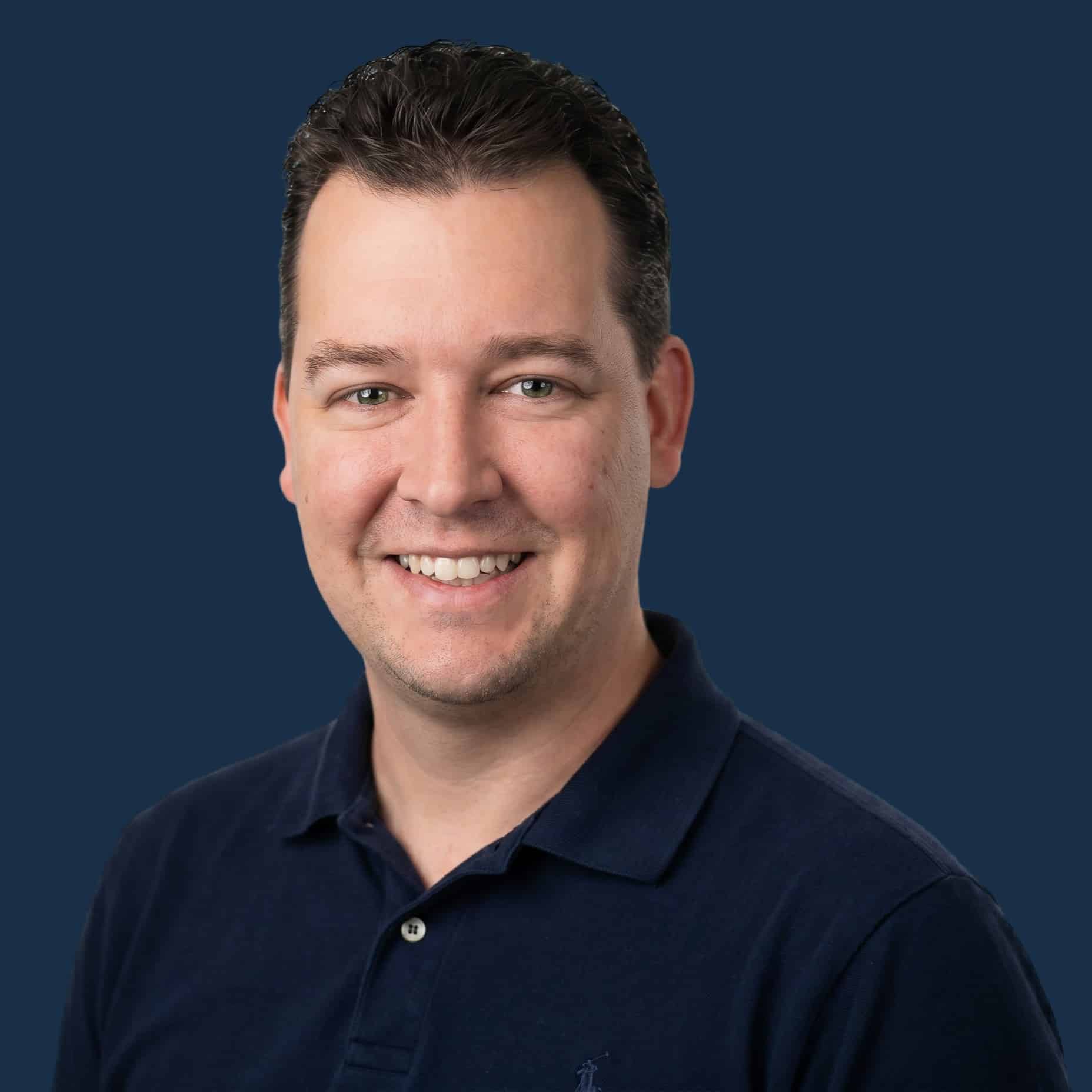 Smiling man with short dark hair wearing a navy blue polo shirt, posed in front of a solid dark blue background.